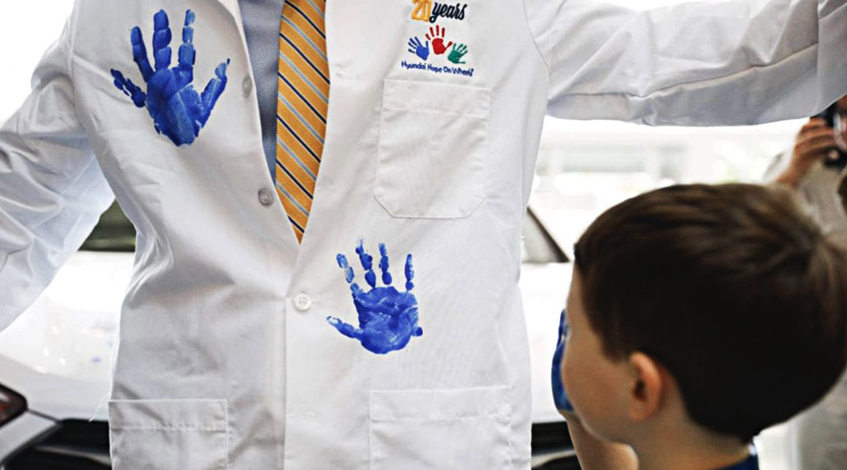Child places a blue handprint on a doctor’s white coat during a Hyundai Hope on Wheels event